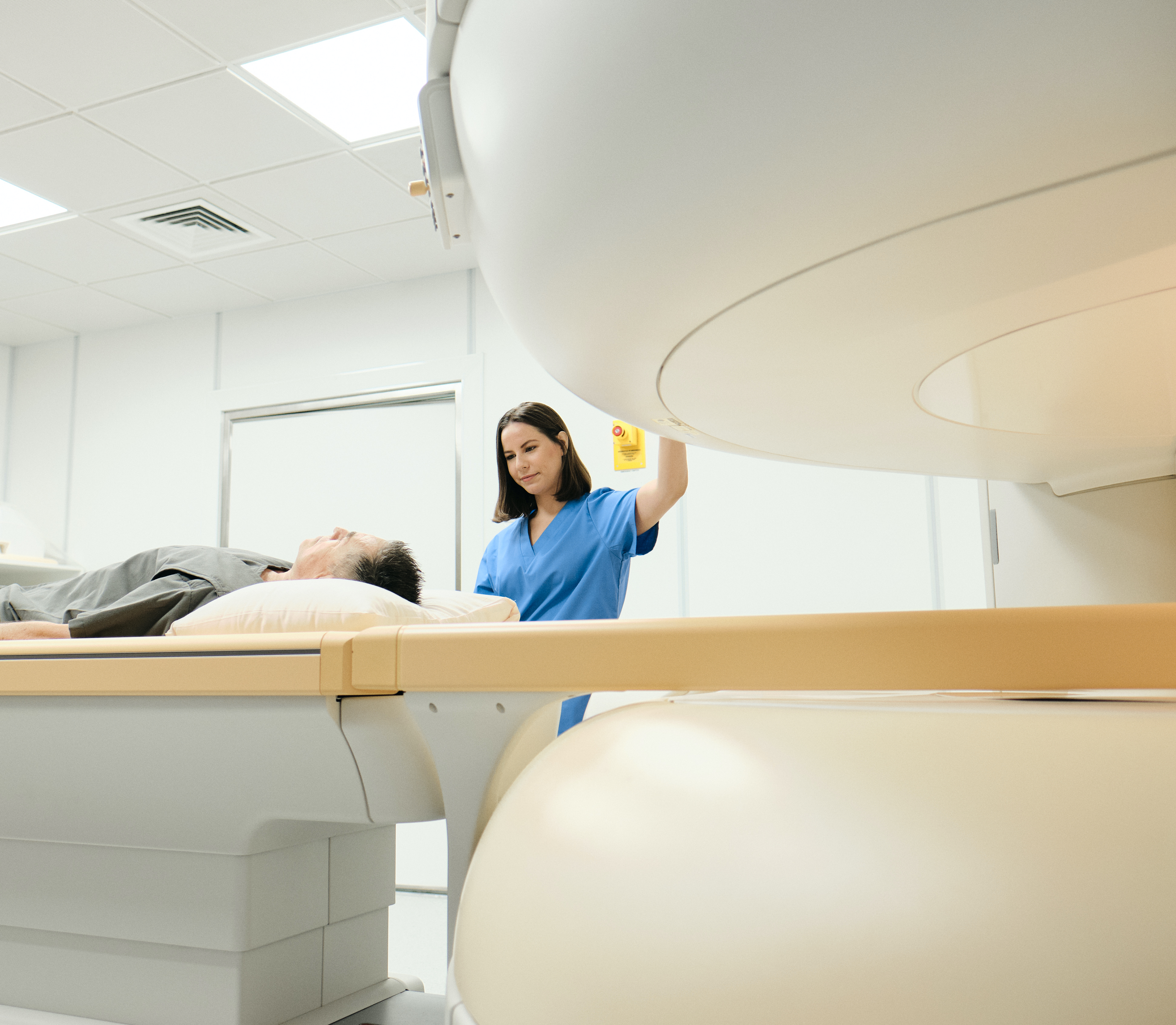 A middle aged patient is entering the MRI scanner. The patient is wearing grey, he is looking at the ceiling. The nurse is wearing blue scrubs, she has lovely brown hair and a friendly smile.