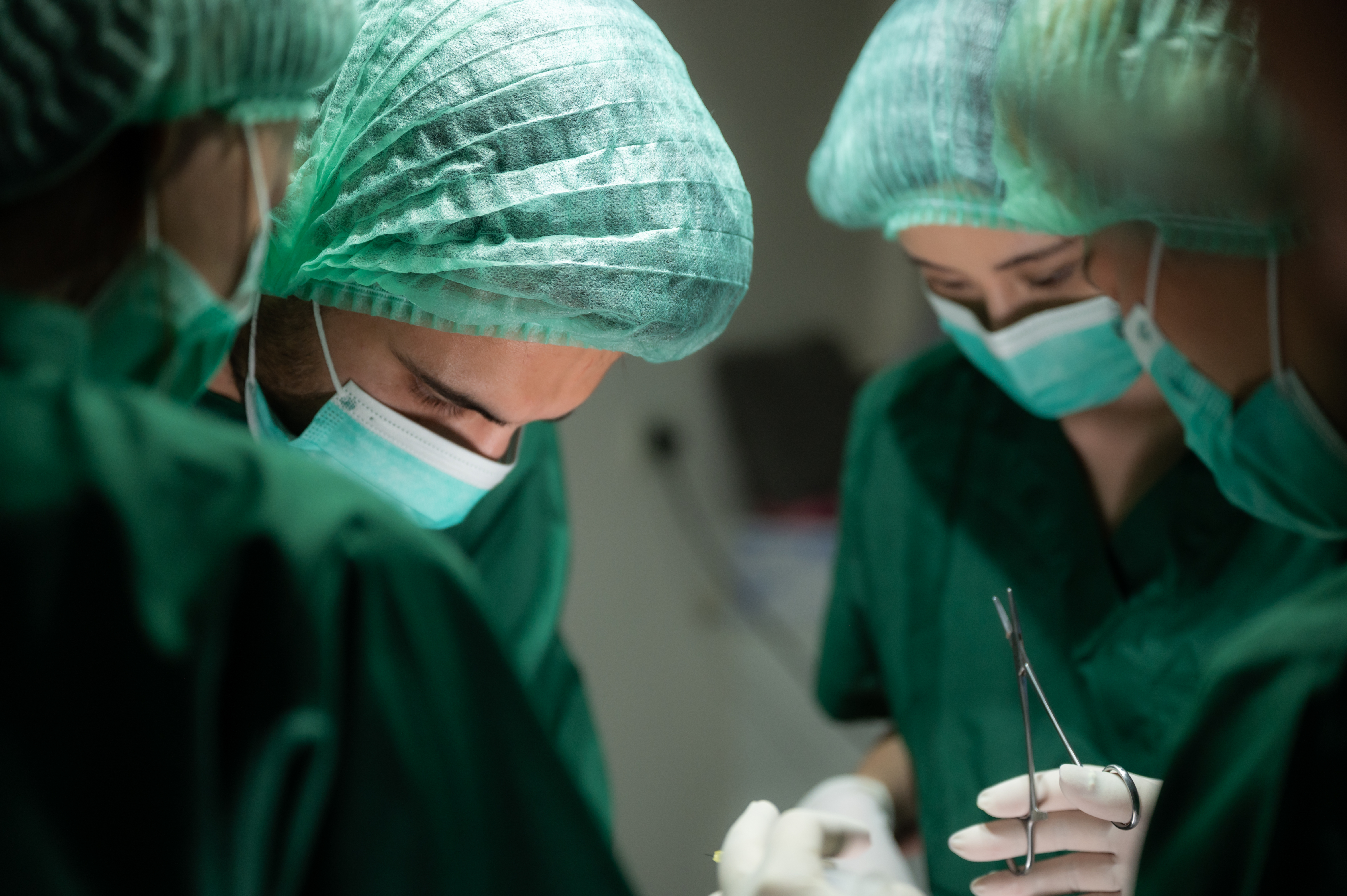 Four surgeons wearing green scrubs and masks and holding surgical tools perform an operation. (Source: envato)